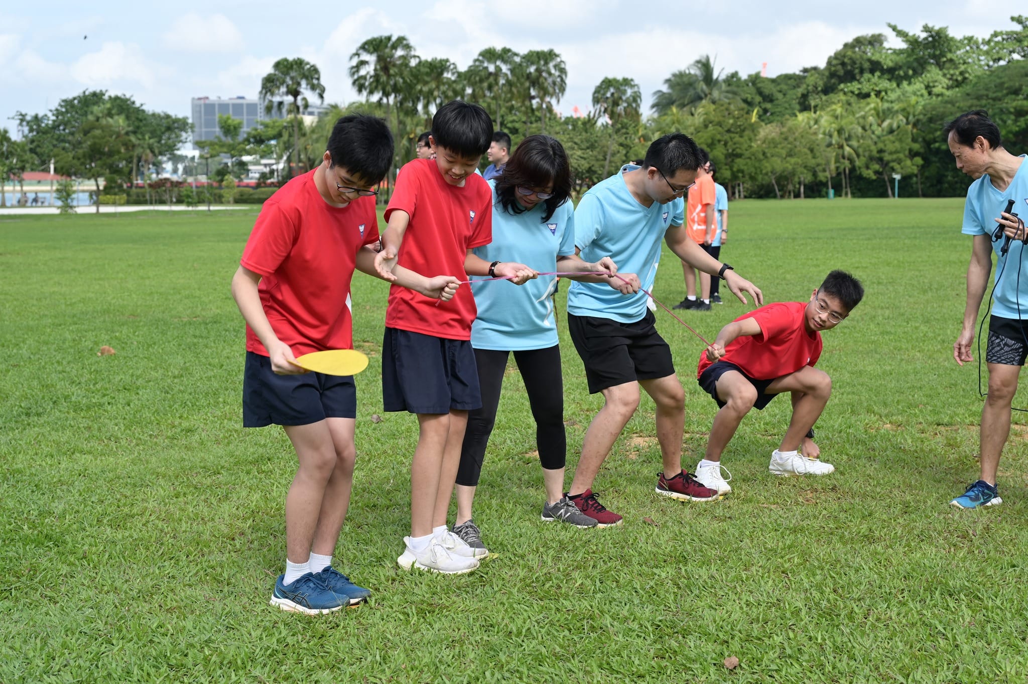 A group of people in red and blue shirts play a team activity on a grassy field, using a rope and yellow disc.